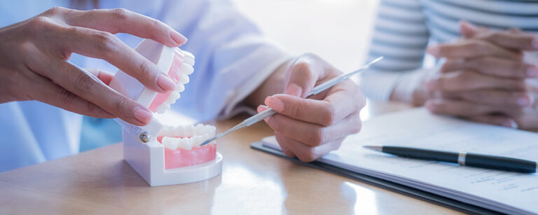 Dentist showing and explaining teeth disease treatment to patient using teeth model denture and explorer mirror tool in dental clinic office. Healthcare concept.